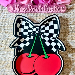 May include: A black and white checkered bow design tops two red cherries with green stems. The design is on a black background. The text "@NurseRondaCreations" is above the design.