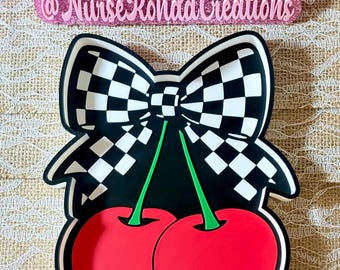 May include: A black and white checkered bow design tops two red cherries with green stems. The design is on a black background. The text "@NurseRondaCreations" is above the design.