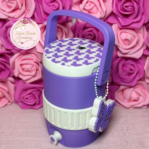 May include: A purple and white water cooler with a handle and a butterfly design on the lid. A white butterfly charm hangs from the side. The cooler has a white band around the middle and a spigot at the bottom.