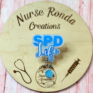 Può includere: Un portabadge azzurro e bianco con la scritta "SPD Life" su sfondo in legno. Il portabadge ha un accento blu glitterato e un supporto in plastica trasparente. Lo sfondo in legno presenta la scritta "Nurse Ronda Creations" e illustrazioni mediche.
