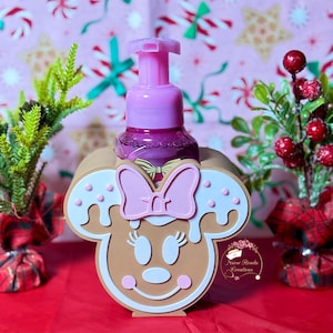 Puede incluir: Un dispensador de jabón rosa con un diseño de pan de jengibre de Minnie Mouse. El dispensador está colocado sobre un fondo festivo con decoraciones navideñas. El dispensador tiene una bomba rosa y un diseño de pan de jengibre marrón y blanco.