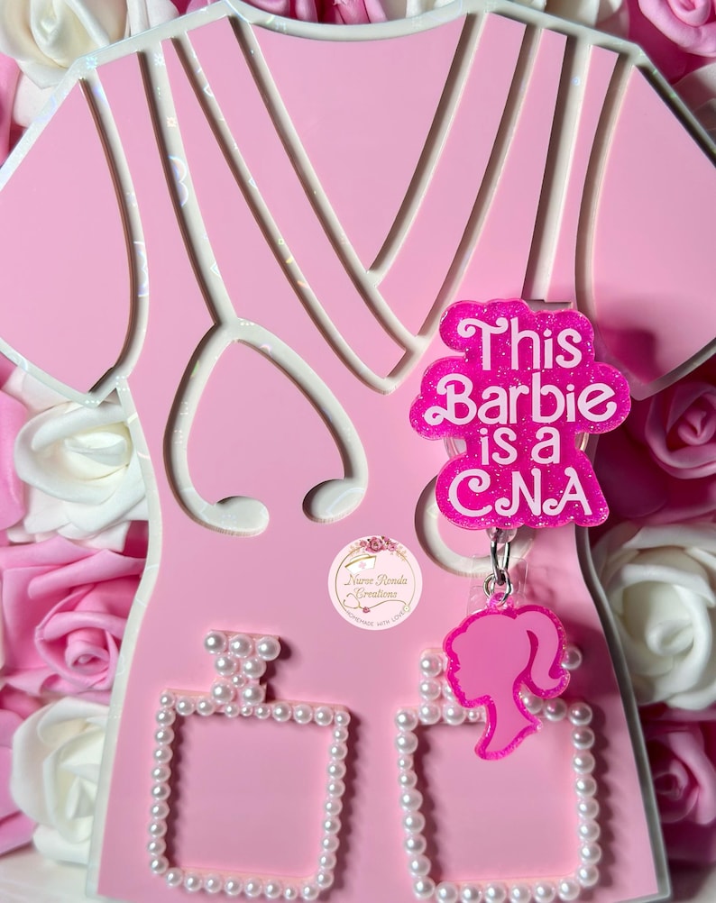 May include: Pink and white cutout of a dress with a pink glitter charm that says "This Barbie is a CNA". The charm is attached to a pink Barbie silhouette charm with a pearl-trimmed frame.