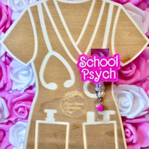 May include: A wooden cutout of a scrub top with a pink glitter badge that says "School Psych". The badge is attached to a retractable badge holder with a pink charm.