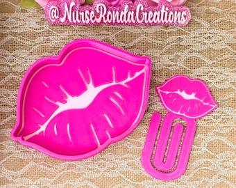 May include: Bright pink lip-shaped cookie cutters. One large lip-shaped cutter and a smaller lip-shaped cutter with a U-shaped handle. The text "@Nurse Ronda Creations" is above the cutters.