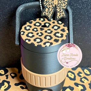 Könnte beinhalten: Schwarzer und beigefarbener isolierter Becher mit Leopardenmusterdeckel und Schleife. Der Becher hat einen Griff und ein beiges Band in der Mitte. Ein Etikett mit dem Text "Nurse Ronda Creations" ist angebracht.
