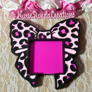 May include: A pink and black bow-shaped tray with a leopard print design. The tray has a rectangular center in a vibrant pink color. The text "@NurseRondaCreations" is above the bow. Decorative pink and white flowers are in the background.