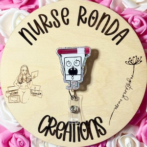 Puede incluir: Un carrete de insignia de brillo blanco y plateado con un personaje de dibujos animados que sostiene una jeringa roja y blanca. El personaje tiene una expresión de sorpresa con ojos grandes y la boca abierta. El texto "Nurse Ronda Creations" está grabado en un fondo de madera.