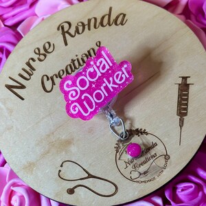 K&ouml;nnte beinhalten: Ein rosa Holz-Kreis mit dem Text "Nurse Ronda Creations" und einem rosa Glitzer-Abzeichen mit der Aufschrift "Social Worker".