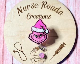 May include: A pink Grinch wearing a Santa hat is attached to a retractable badge holder. The badge holder has a clear plastic cover with a pink glitter circle and the text "Nurse Creations" printed on it. The badge holder is on a wooden background with a stethoscope and a syringe.