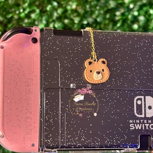 May include: A pink and black Nintendo Switch console with a clear glitter case. A gold chain with a brown bear charm hangs from the top of the case. The text "Nurse Rondia Creations" is printed on the case.