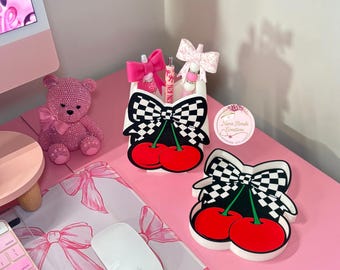 May include: A pink desk setup featuring a pink keyboard, mouse, and a decorative pink teddy bear. The desk also has a white container with a black and white checkered bow and red cherries design. There is also a matching coaster.