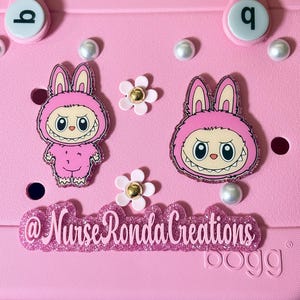 May include: Pink bag with decorative charms. The charms include two pink bunny figures with large eyes and a toothy grin, white flower charms with gold centers, and faux pearl accents. The text "@Nurse Ronda Creations" is also visible.