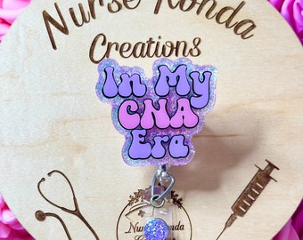 May include: A wooden circle with the text "Nurse Ronda Creations" and a purple and pink glitter badge holder with the text "In My CNA Era". The badge holder is attached to a retractable badge reel. A stethoscope and a syringe are engraved on the wood.