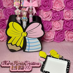 May include: Decorative pens with bows and character figures in a pencil-shaped holder. A matching tray and logo are also visible, set against a backdrop of pink roses. The items are in a pink and black color scheme.