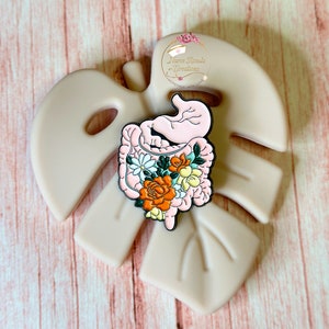 May include: A pink and black enamel pin featuring a stylized human intestine with a floral arrangement of orange, yellow, and white flowers. The pin is on a light brown background.