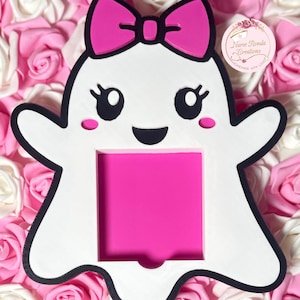 May include: A white ghost-shaped picture frame with a pink bow, black outline, and a pink interior. The frame has a smiling face with black eyes and a black mouth. The background is a mix of pink and white roses.