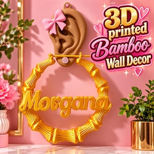 Op de afbeelding: Een 3D-geprinte wanddecoratie met een bruine, oorvormige houder met een roze strik, waaraan een grote goudkleurige bamboe-stijl oorring hangt. De naam Morgana staat in gouden letters op de ring. Tekst: 3D printed Bamboo Wall Decor.