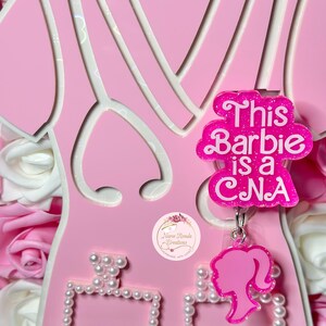 May include: Pink Barbie doll cutout with a pink and white dress and a pink charm that says "This Barbie is a CNA". The doll is holding a pink perfume bottle with a pearl trim.