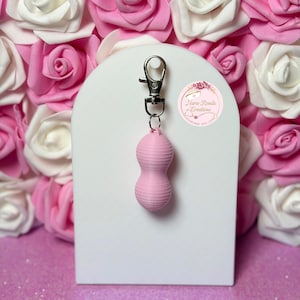 Könnte beinhalten: Ein rosa, stundenförmiger Schlüsselanhänger mit einem silberfarbenen Verschluss. Der Anhänger hat eine gerippte Textur und hängt vor einem weißen Bogen. Der Hintergrund zeigt rosa und weiße Rosen.
