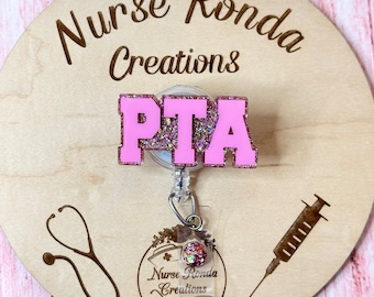 Puede incluir: Carrete de insignia de brillo rosa con las letras PTA en rosa con contornos blancos. El carrete está unido a un porta-insignias de plástico transparente con un estetoscopio y un diseño de jeringa sobre un fondo de madera. El texto "Nurse Ronda Creations" también está en el fondo.