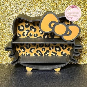 May include: A black, three-tiered shelf shaped like a cartoon cat's head. The interior has a leopard print design. The bow is yellow with a black outline. The text "Nurse Ronda Creations" is visible on a pink circular sticker.
