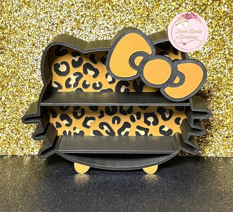 May include: A black, three-tiered shelf shaped like a cartoon cat's head. The interior has a leopard print design. The bow is yellow with a black outline. The text "Nurse Ronda Creations" is visible on a pink circular sticker.
