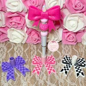 May include: A collection of decorative pens with bow accents in pink, purple, and black and white checkered patterns. The pens have a pink bow at the top and a bedazzled bead. The image includes the text "Nurse Renda Creations".