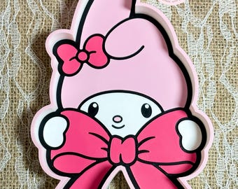 May include: A pink My Melody-shaped tray with a large bow and a smaller bow on the head. The tray has a black outline and a white face. The tray is a cute and decorative item.