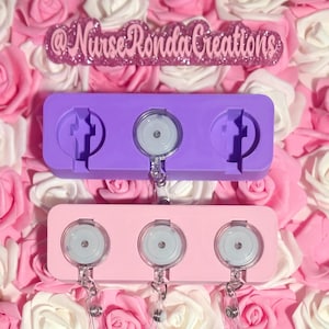 Op de afbeelding: Een close-up van twee badge-rolhouders. De bovenste houder is paars en de onderste houder is roze. Elke houder heeft drie ronde badge-rollen met helder plastic en zilveren accenten. De achtergrond is een bed van roze en witte rozen, met de tekst "@Nurse Ronda Creations".