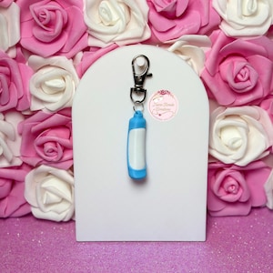 May include: A blue and white keychain with a silver clasp hangs in front of a white arch. The background features pink and white roses. The keychain has a cylindrical design with a white center and blue accents. The image also includes a small sticker with text.
