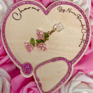 May include: A wooden heart-shaped display with a stethoscope design and the text "Charms By Nurse Ronda Creations." The display features a pink glitter border and a charm with pink flower and green leaf accents.