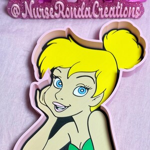 May include: A cookie cutter in the shape of Tinkerbell, featuring yellow hair, blue eyes, and a green top. The design is outlined in black and set within a pink frame. Pink roses and the text "Nurse Ronda Creations" are in the background.
