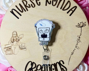 May include: A white and blue glitter badge reel with a cartoon character with a surprised expression. The reel is attached to a clear plastic retractable badge holder. The background is a wooden circle with the text "Nurse Ronda Creations" written on it.
