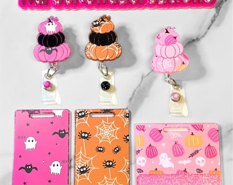 May include: Three Halloween themed retractable badge holders with different designs. The first badge holder features a stack of pink pumpkins with a ghost and bats. The second badge holder features a stack of orange pumpkins with a spider web and spiders. The third badge holder features a stack of pink pumpkins with a ghost, bats, and skulls. Each badge holder has a glitter bottom.