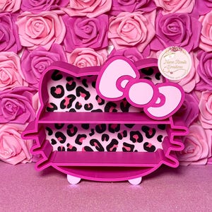 May include: A pink cat-shaped shelf with three tiers and a bow detail. The interior has a leopard print pattern. The shelf is displayed against a backdrop of pink and rose-coloured paper roses.