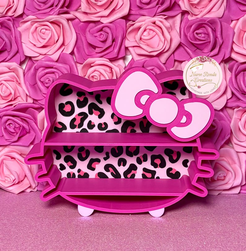 May include: A pink cat-shaped shelf with three tiers and a bow detail. The interior has a leopard print pattern. The shelf is displayed against a backdrop of pink and rose-coloured paper roses.