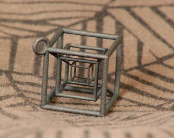 May include: A silver-colored metal pendant in the shape of a cube with a smaller cube inside. The cube is made of wire and has a loop at the top for hanging.