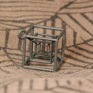 May include: A silver-colored metal pendant in the shape of a cube with a smaller cube inside. The cube is made of wire and has a loop at the top for hanging.