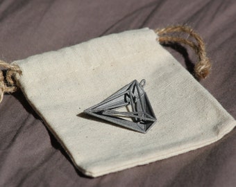 May include: A grey, three-dimensional geometric pendant with a hollow, angular frame structure, resting on a small, off-white, rectangular fabric drawstring pouch made of woven material with natural fiber cords at the top closure.