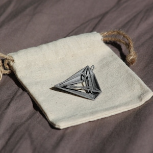 May include: A grey, three-dimensional geometric pendant with a hollow, angular frame structure, resting on a small, off-white, rectangular fabric drawstring pouch made of woven material with natural fiber cords at the top closure.