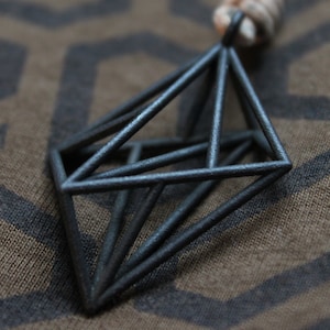 May include: A matte black geometric pendant featuring a three-dimensional wireframe structure shaped like a double pyramid. The pendant is suspended from a light-colored cord and displays a textured, dark metallic finish with sharp, clean lines.