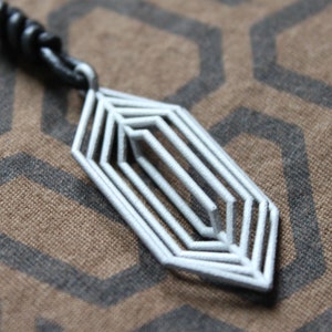 May include: A silver geometric pendant necklace with a black leather cord. The pendant is a multi-layered, abstract design with a pointed shape.