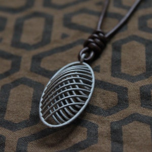 May include: A silver pendant necklace with a geometric design hangs from a brown leather cord. The pendant is oval shaped and features a series of intersecting lines creating a grid pattern.