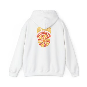 Puede incluir: Sudadera con capucha blanca con un estampado gráfico de un sol con el texto "Keep Chasing Sunsets" en amarillo y naranja.