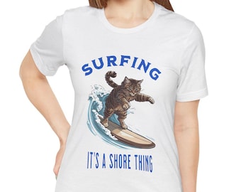 Puede incluir: Camiseta blanca con un gráfico azul de un gato surfeando en una ola. El texto "SURFING" está por encima del gato y "IT'S A SHORE THING" está por debajo del gato.