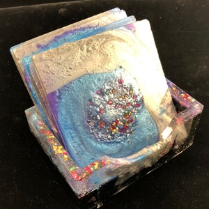 Op de afbeelding: Set van vier blauwe, zilveren en paarse onderzetters met iriserende glitter ingebed in de hars. De onderzetters zijn gestapeld en zitten in een transparante harshouder.