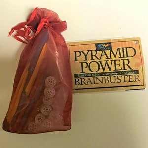 May include: A red mesh bag filled with colorful wooden sticks and white plastic spheres. The bag is next to a card that reads "Pyramid Power Brainbuster" with the text "Can you solve the mystery of the ages?"