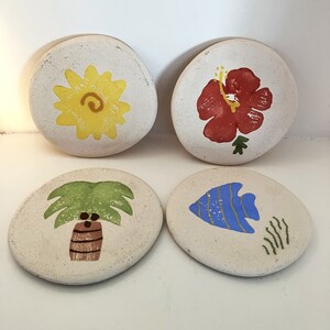 Op de afbeelding: Vier witte keramische onderzetters met verschillende ontwerpen. Een onderzetter heeft een gele bloem, een andere een rode hibiscus, een derde een groene palmboom en de laatste een blauwe vis.