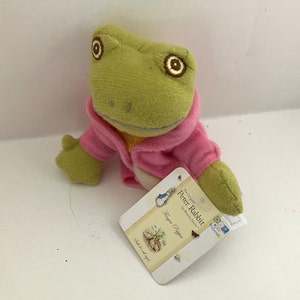 May include: A green frog wearing a pink jacket, a finger puppet from the Peter Rabbit collection by Beatrix Potter. The tag reads "The Original Peter Rabbit by Beatrix Potter Finger Puppet".