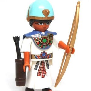 May include: A toy figure of an Egyptian pharaoh, wearing a blue helmet and white robe with colorful accents. The figure holds a golden bow and has a quiver with arrows. The figure is a collectible toy.